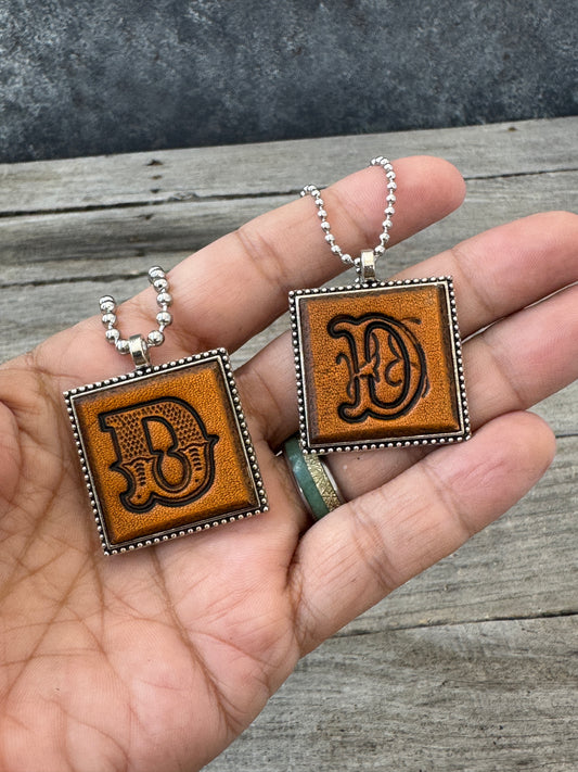 Western Block and Floral Script letter D leather pendants held in hand to show their 1-inch size
