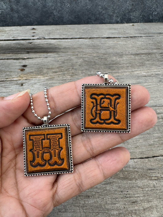 Two Letter H leather initial necklaces held in hand showing the approximately one-inch square pendants in Western Block and Floral Script styles.