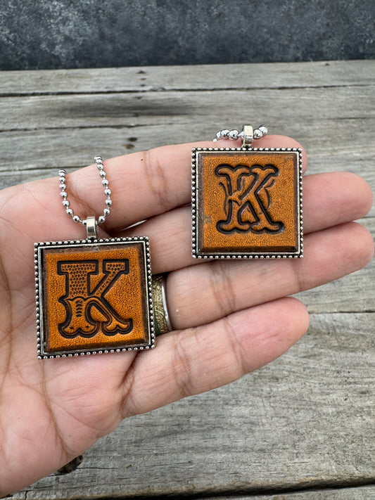 Western Block and Floral Script Letter K leather initial necklaces held in hand to show the size of the square silver-tone pendants.