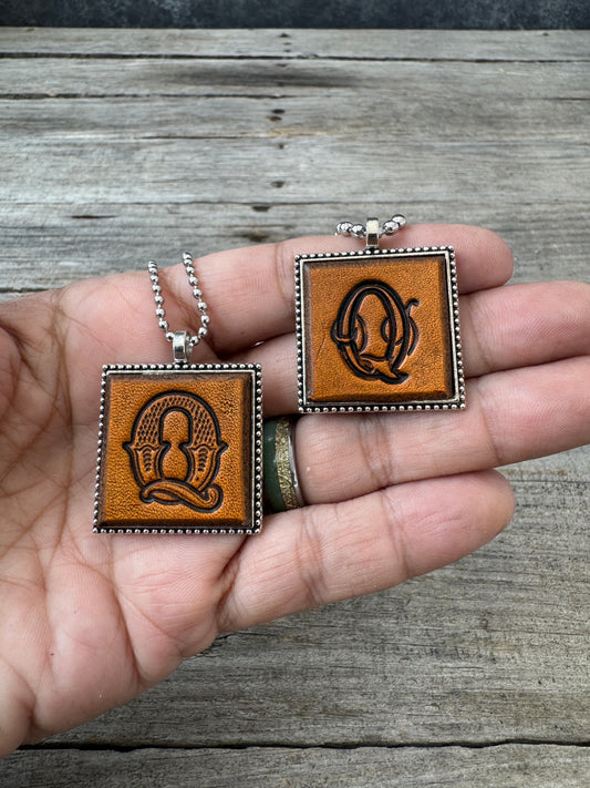 Western Block and Floral Script Letter Q leather necklaces held in hand for size comparison
