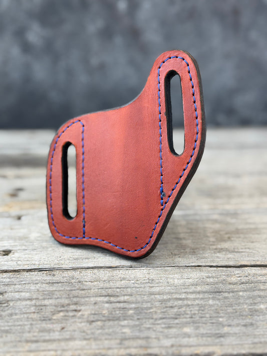 A brown leather knife sheath with blue contrast stitching, standing upright on a rustic wooden surface against a dark gray background.