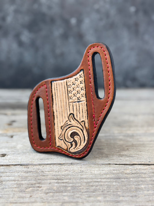 A brown leather knife sheath with a hand-carved wooden American flag and floral emblem on the front, displayed on a rustic wooden surface with a blurred gray background.