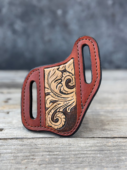 A custom leather knife sheath with a tooled floral design on the front, resting on a rustic wooden surface with a blurred gray background.