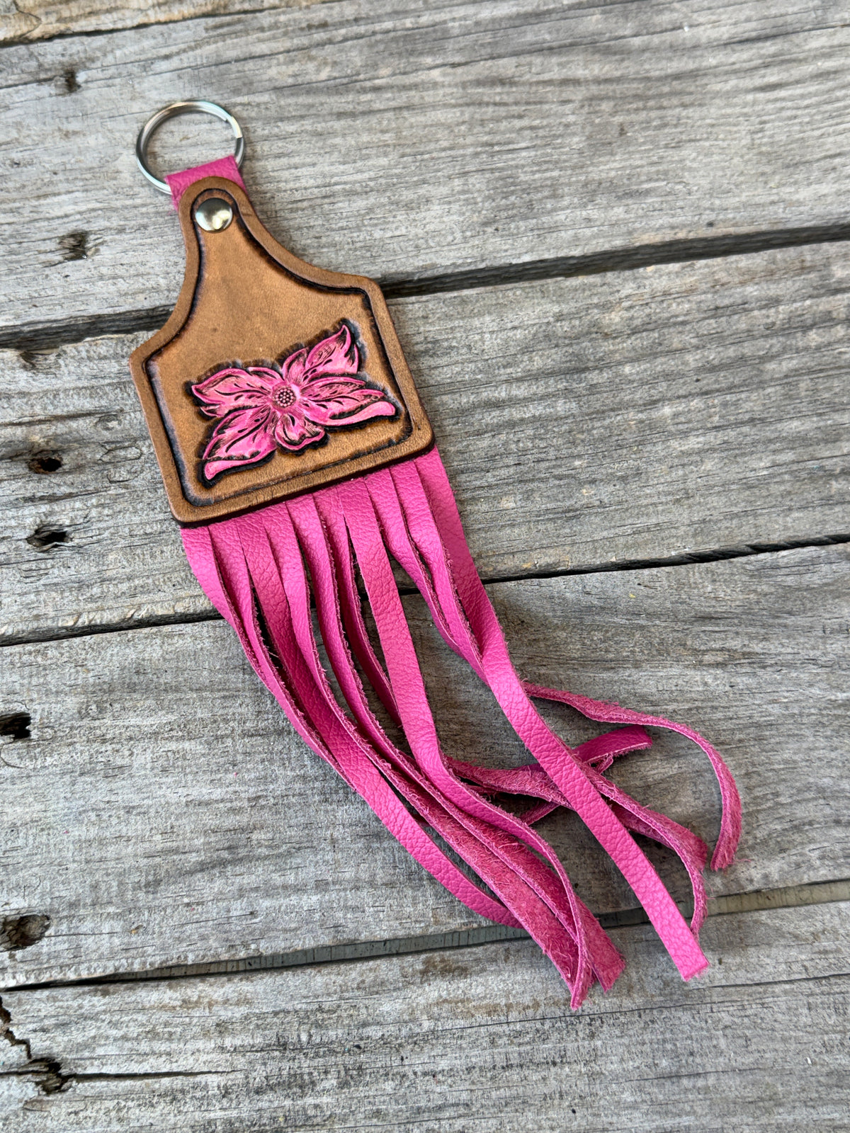 A pink leather keychain with a carved flower design and long fringe, resting on a weathered gray wooden surface.