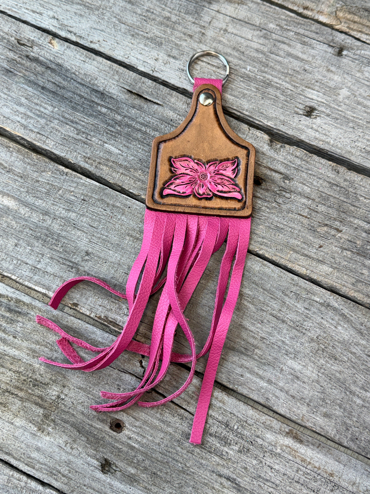 A pink and brown leather keychain featuring a carved flower design and long fringe, displayed on a weathered gray wooden surface.