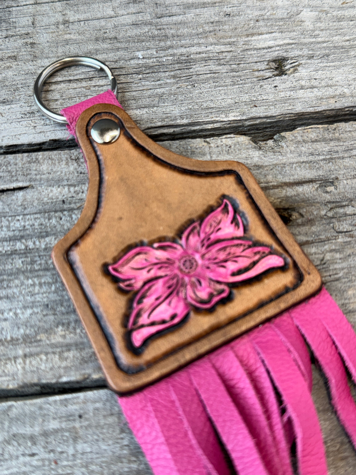 A pink and brown leather keychain featuring a carved pink flower on a brown leather tag, with bright pink fringe and a silver split ring, resting on a rustic gray wooden surface.