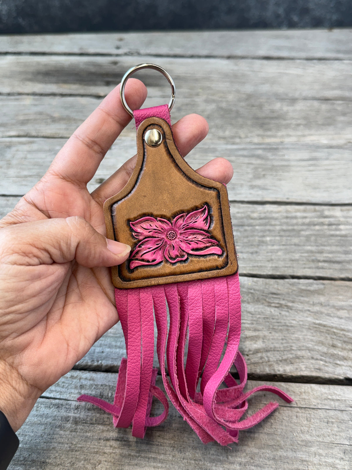 A hand holding a pink and brown leather keychain with a floral design and long fringe, set against a rustic wooden plank background.