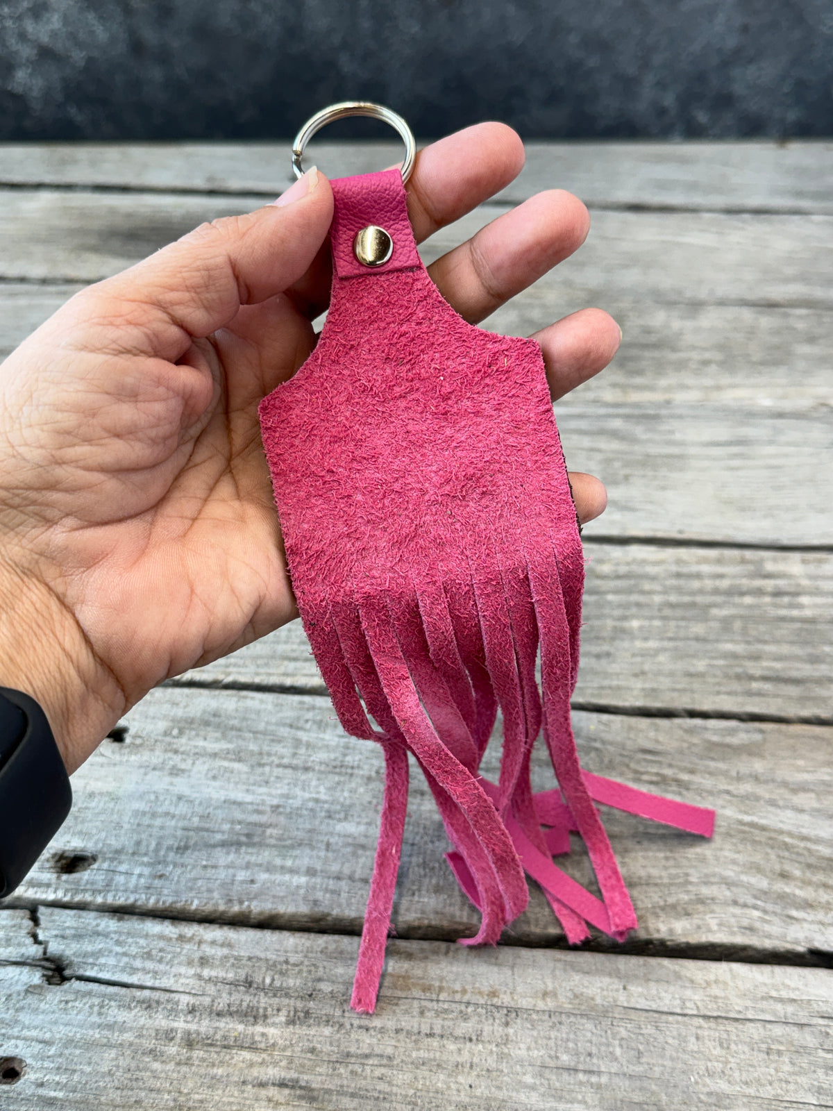 A hand holding a bright pink faux-leather fringe keychain with a silver metal key ring, set against a rustic gray wooden plank background.