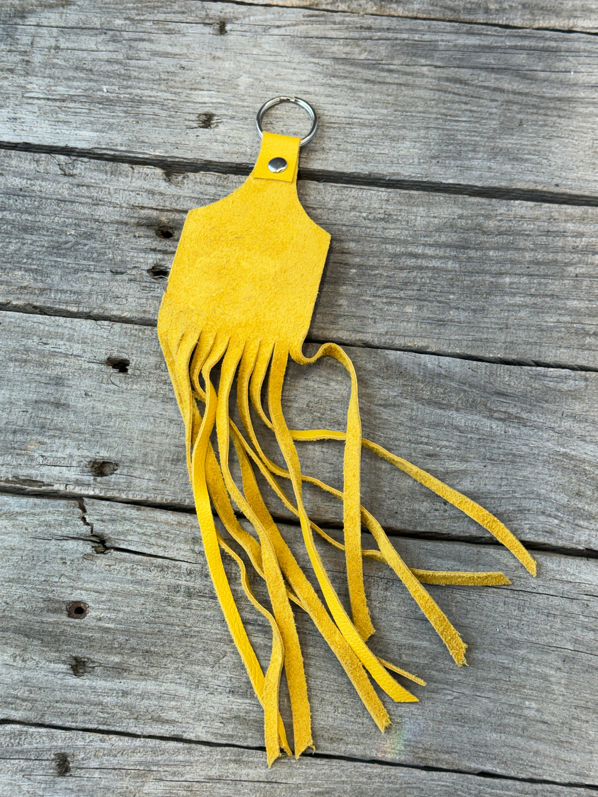 A bright yellow suede fringe keychain with a silver metal key ring, displayed on a weathered gray wooden plank surface.