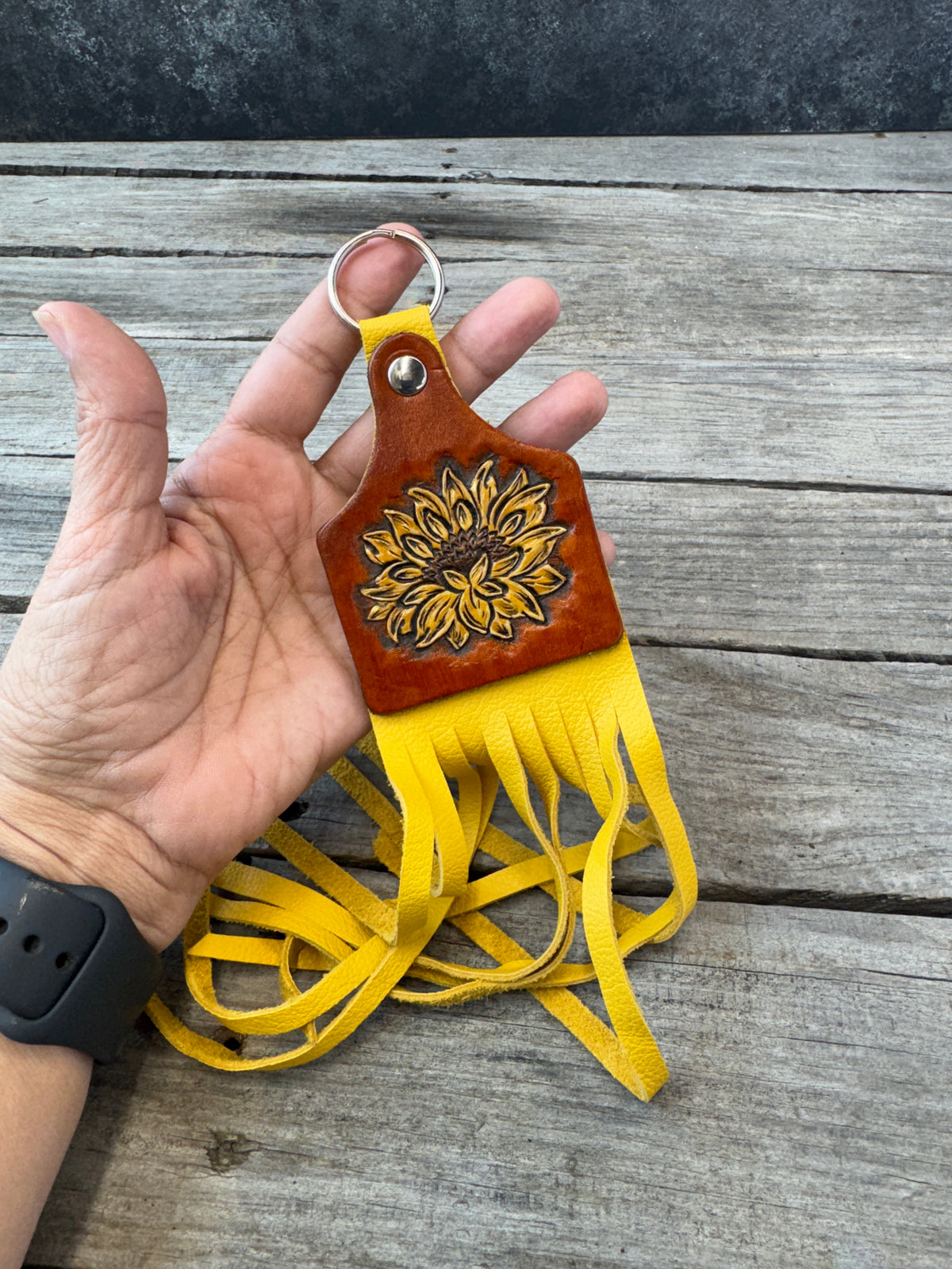 A hand holds a brown leather keychain with a yellow sunflower design and long yellow fringe, set against a rustic gray wooden background.