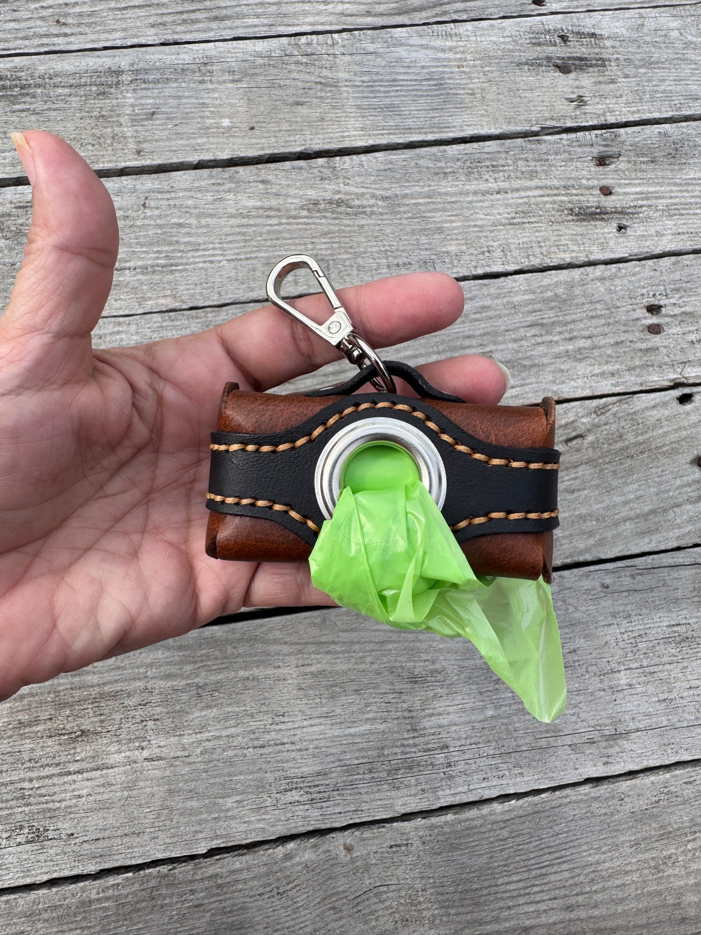A hand holding a small brown leather dog poop bag holder with a silver clasp and a bright green plastic bag protruding from the front, set against a gray wooden plank background.
