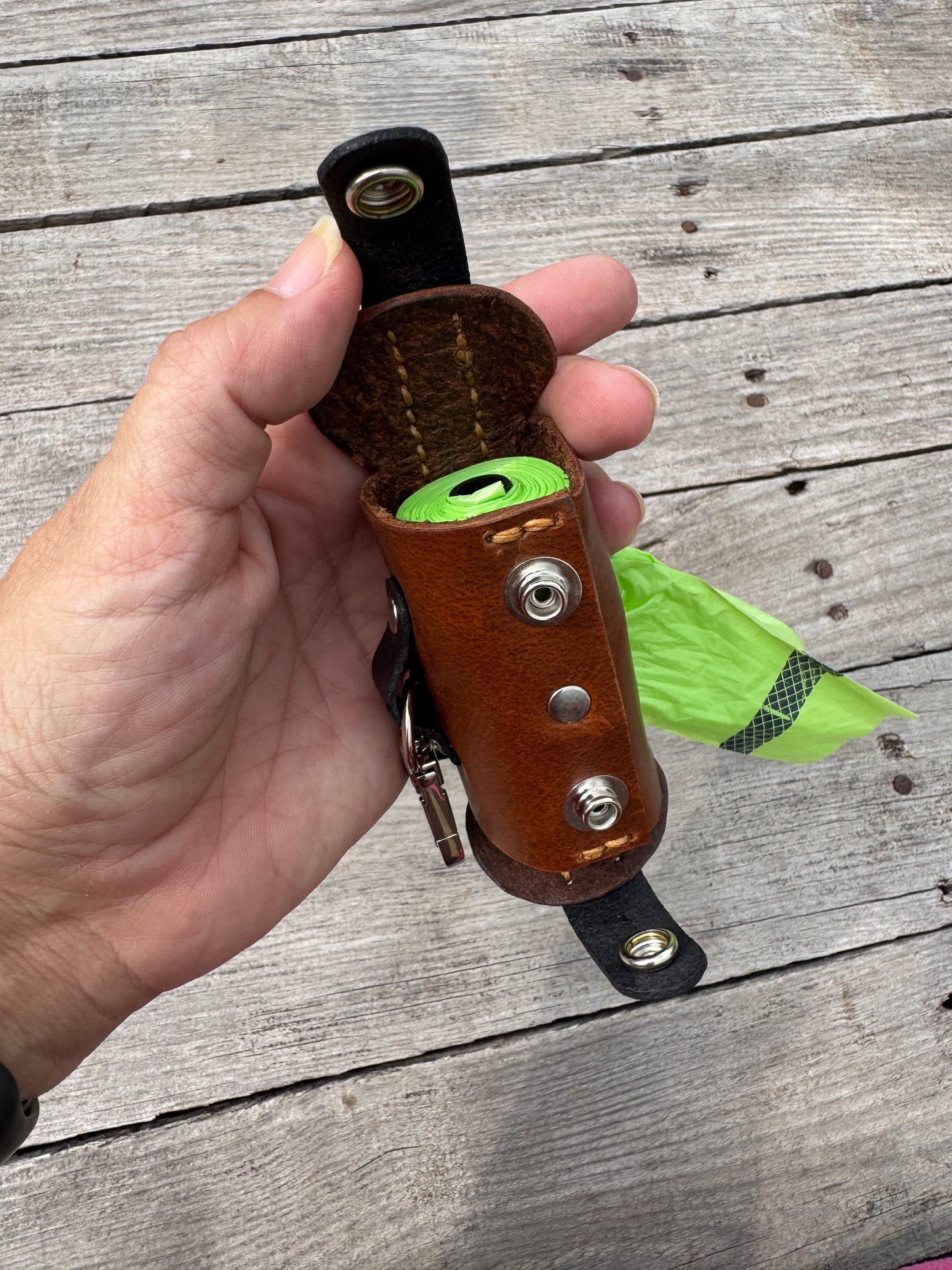 A hand holding a brown leather dog leash holder with a green rolled leash and a bright green bag, set against a weathered gray wooden plank background.
