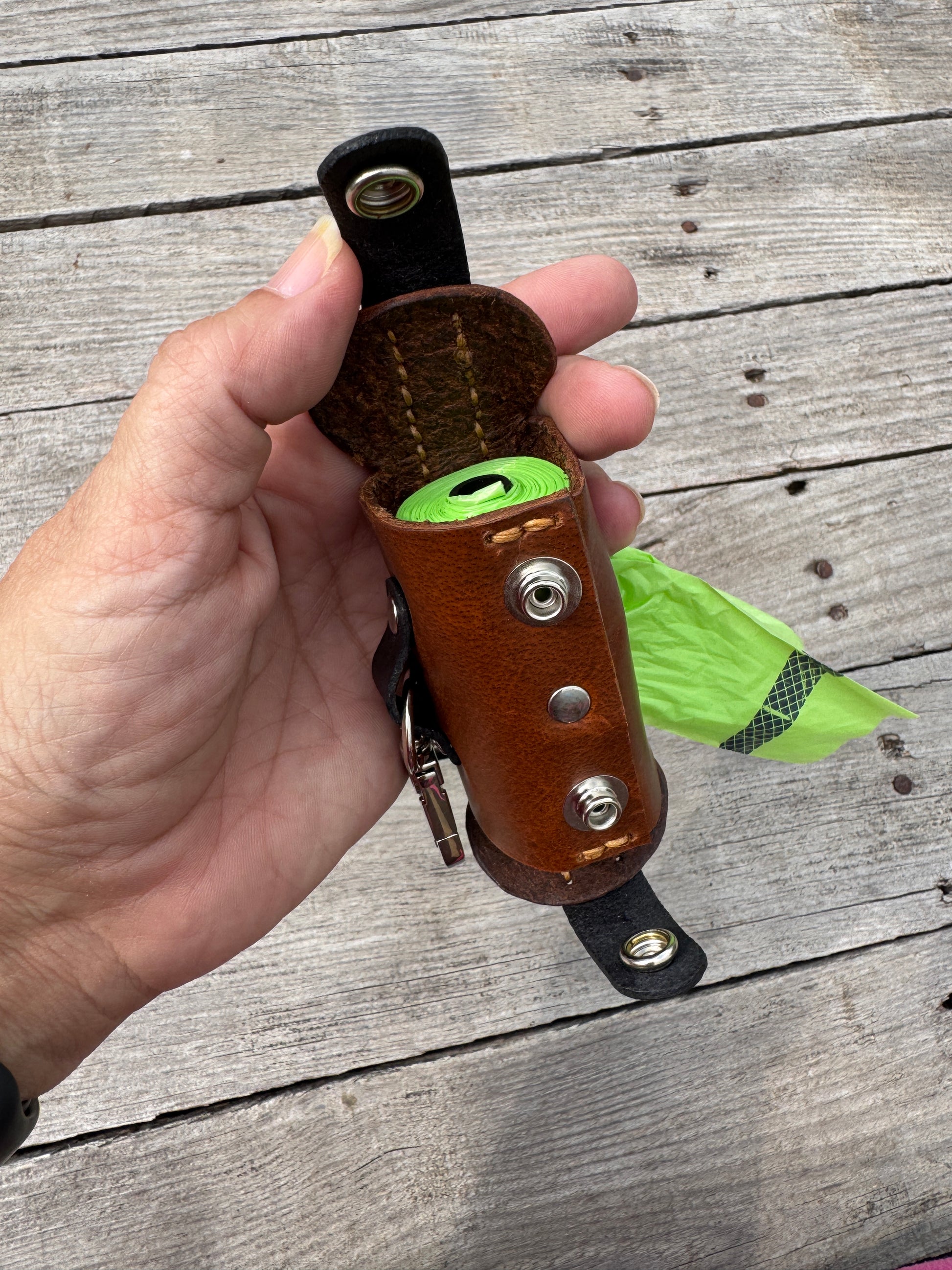 A hand holding a brown leather dog leash holder with a green rolled leash and a bright green bag, set against a weathered gray wooden plank background.