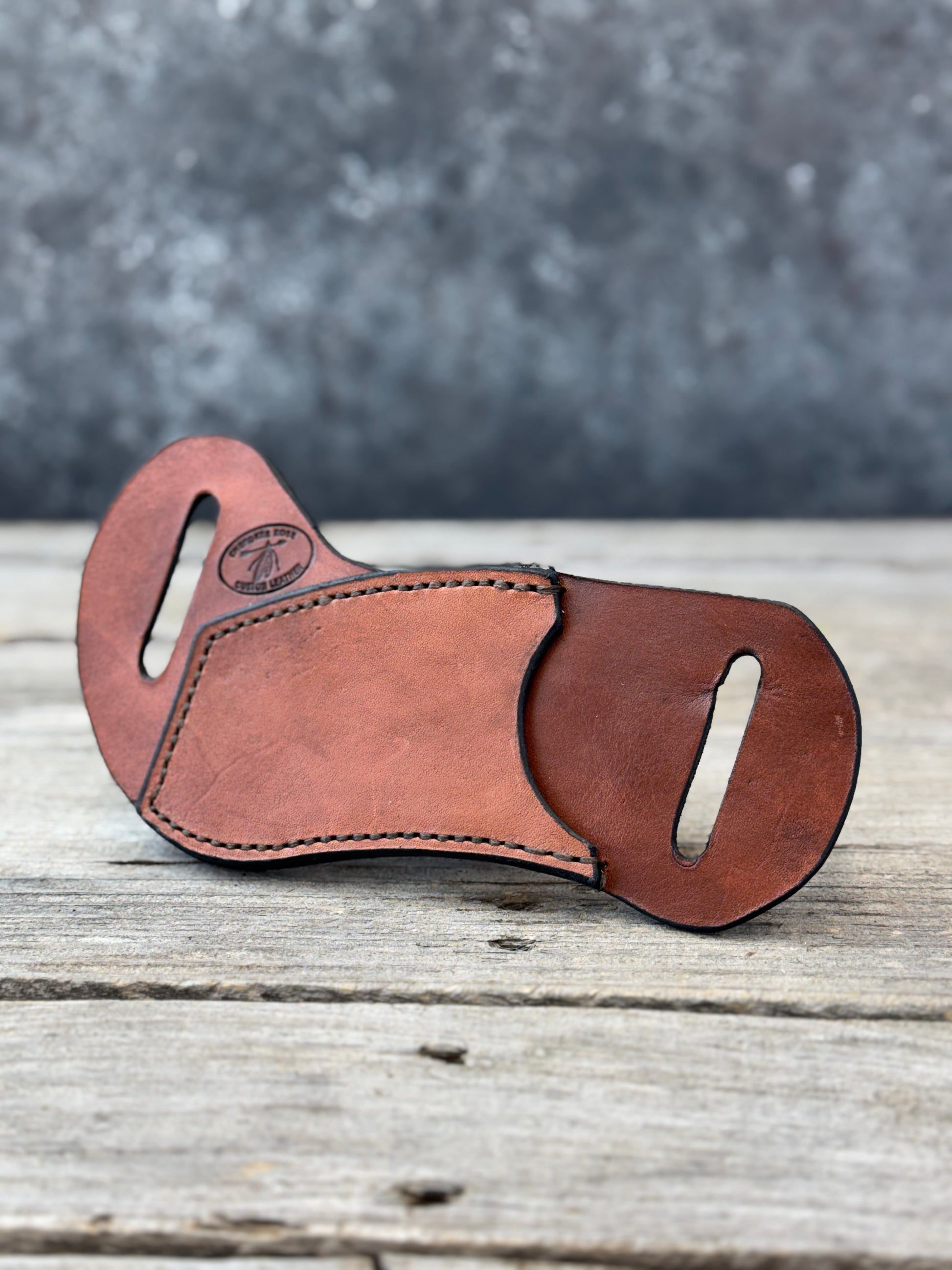 A brown leather multi-tool sheath with two belt cutouts, resting on a rustic wooden surface with a textured gray background.