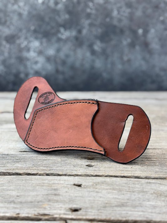 A brown leather multi-tool sheath with two belt cutouts, resting on a rustic wooden surface with a textured gray background.