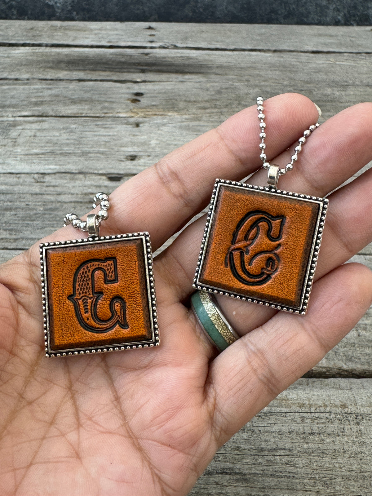 Western Block and Floral Script letter C leather pendants held in hand to show their 1-inch size