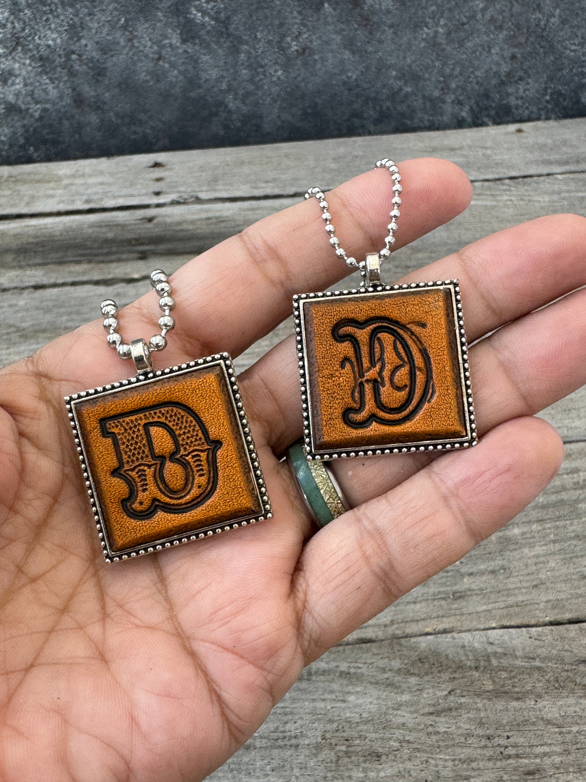 Western Block and Floral Script letter D leather pendants held in hand to show their 1-inch size