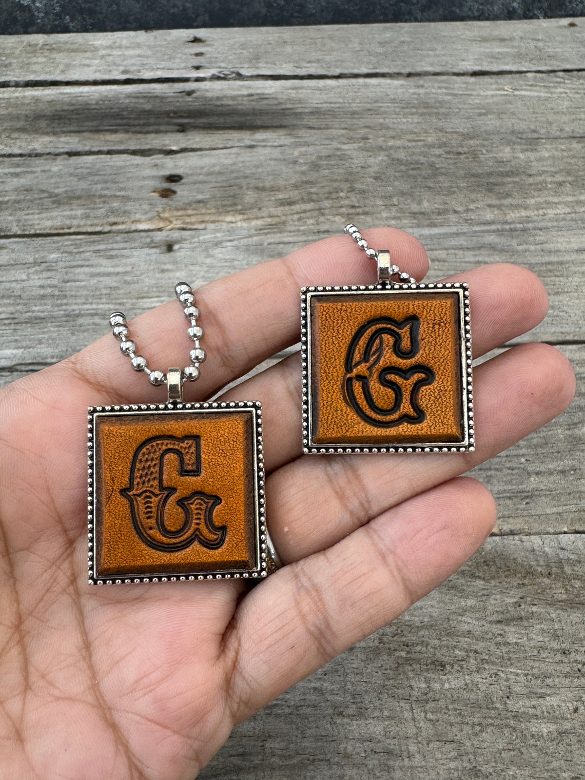 Western Block and Floral Script letter G leather pendants held in hand to show their 1-inch size