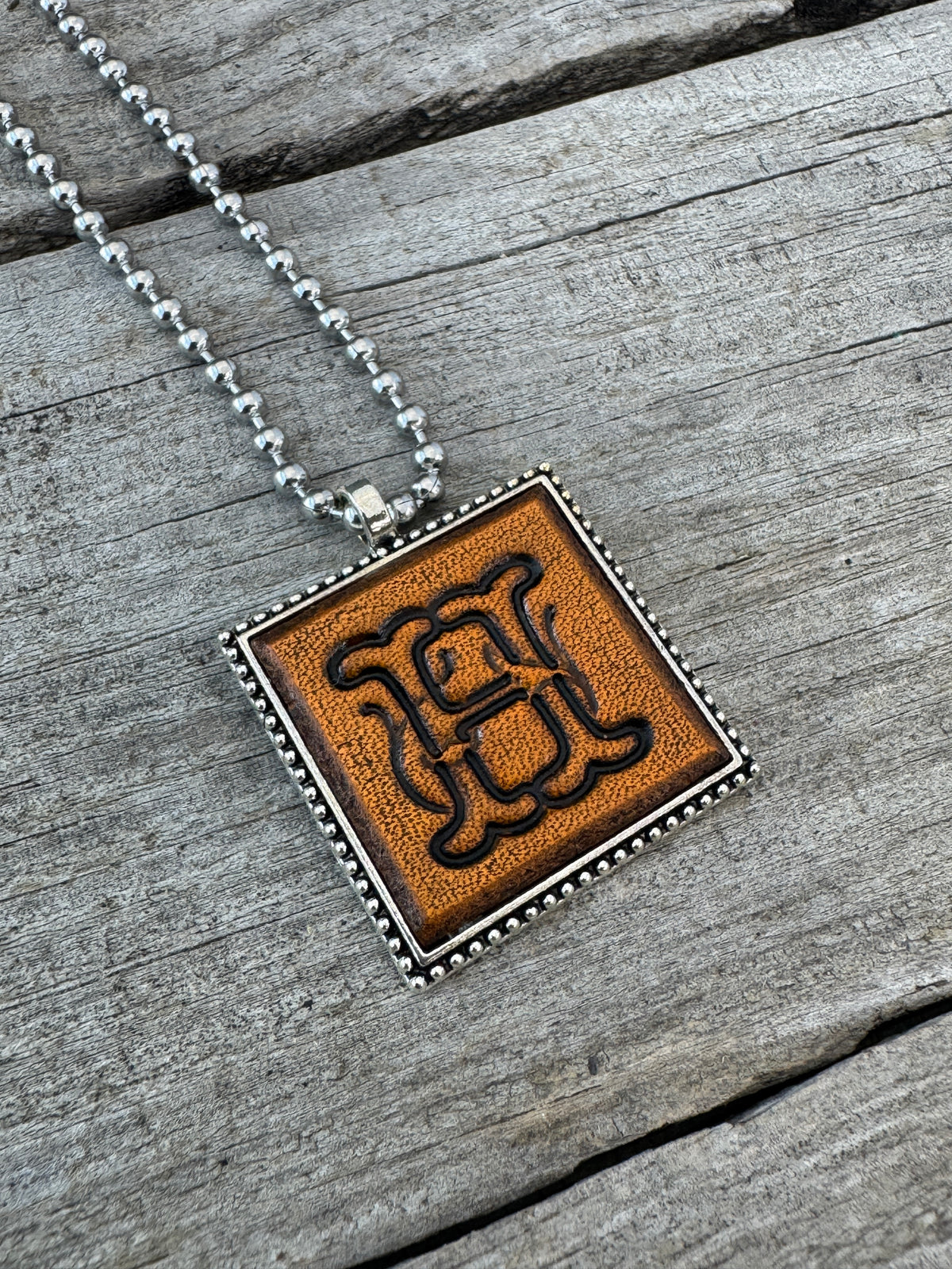 Handmade Letter H leather initial necklace featuring elegant Floral Script lettering in a decorative silver-tone square pendant on rustic wood.