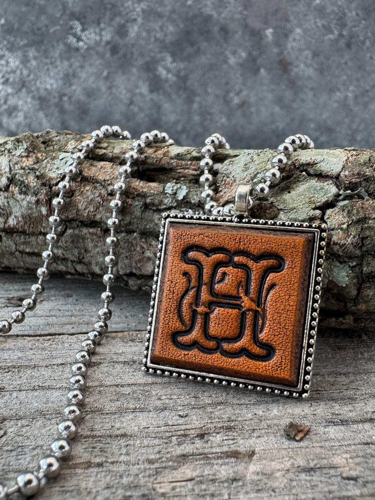Floral Script Letter H leather pendant necklace with silver-tone ball chain displayed against rustic wood and natural bark.