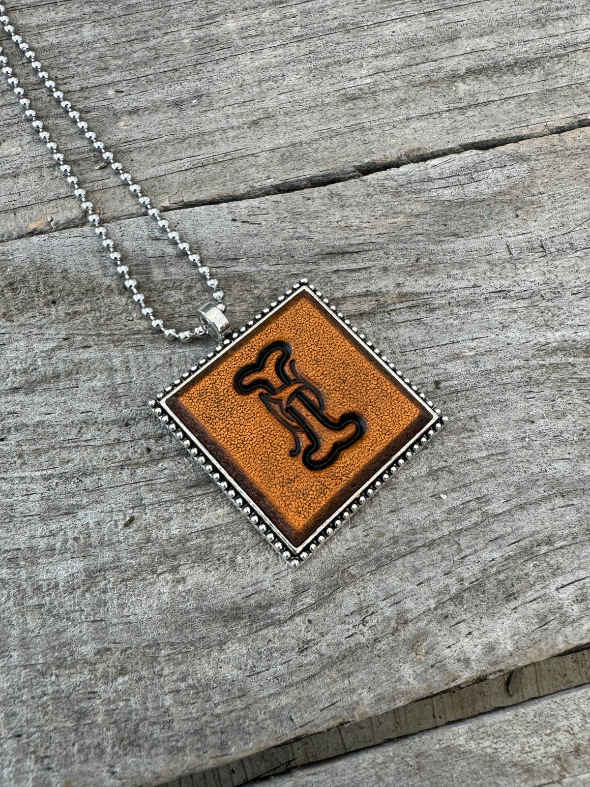Handcrafted Letter I leather initial necklace featuring elegant Floral Script lettering in a decorative silver-tone square pendant on rustic wood.