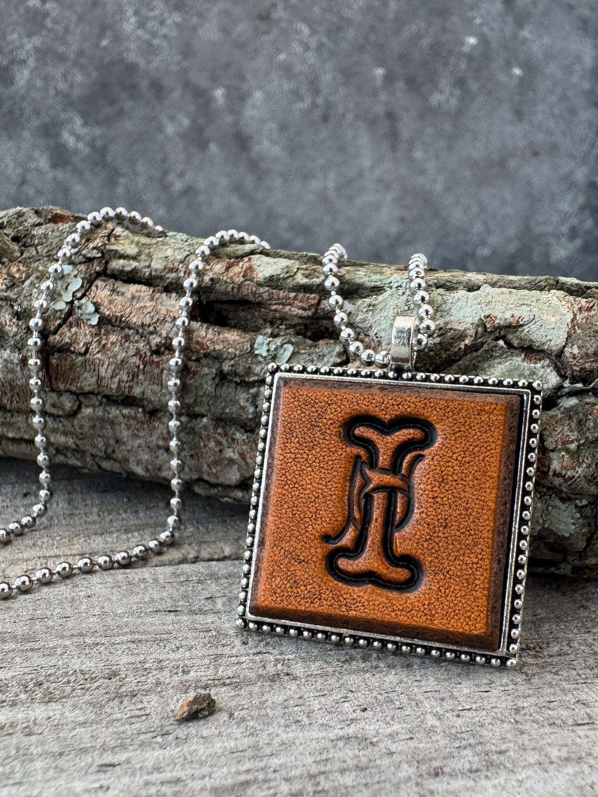 Floral Script Letter I leather pendant necklace with silver-tone ball chain displayed against natural tree bark and weathered wood.