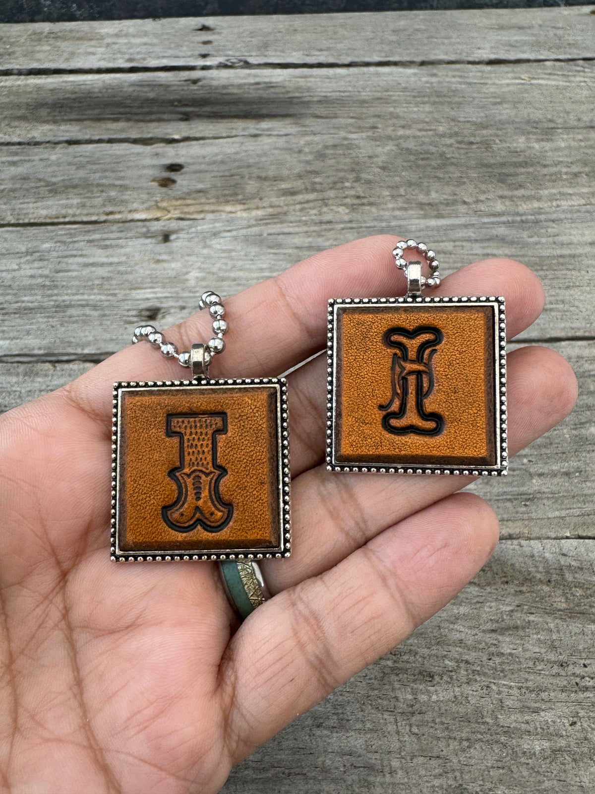 Western Block and Floral Script Letter I leather initial necklaces held in hand to show the size of the square silver-tone pendants.