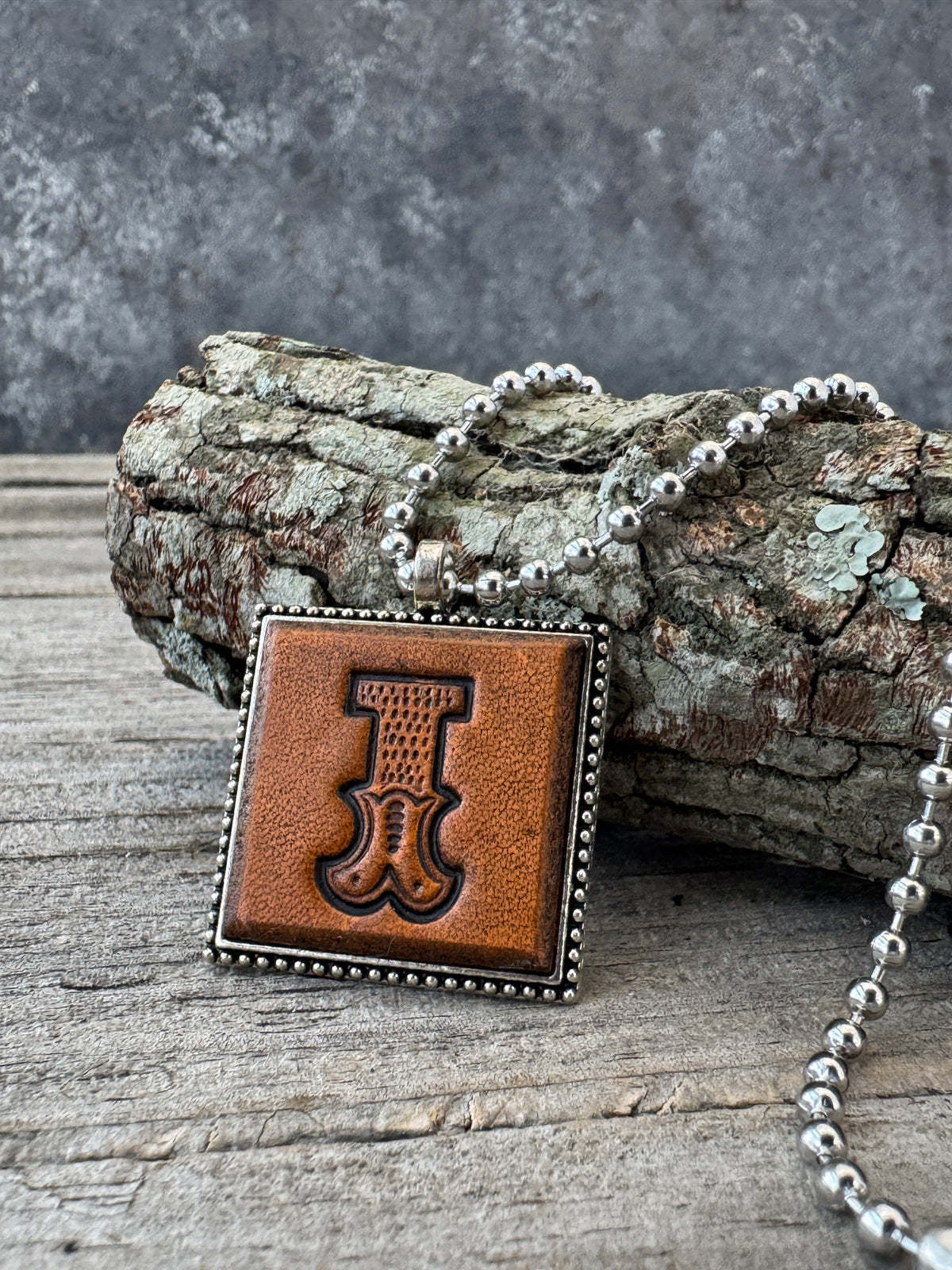 Hand-stamped Western Block Letter I leather necklace with decorative silver-tone frame and ball chain displayed against rustic tree bark.