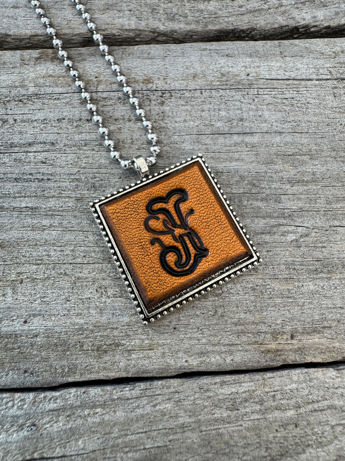 Handcrafted Letter J leather initial necklace featuring elegant Floral Script lettering stamped into warm brown leather with a decorative silver-tone frame.