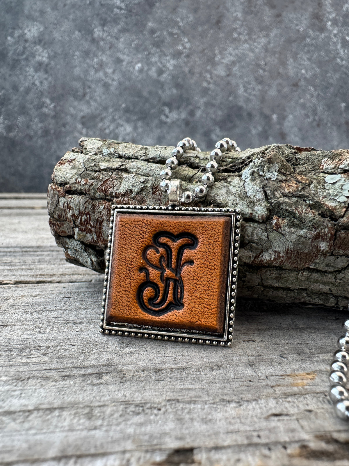 Floral Script Letter J leather pendant necklace with silver-tone ball chain displayed against natural tree bark and rustic weathered wood.