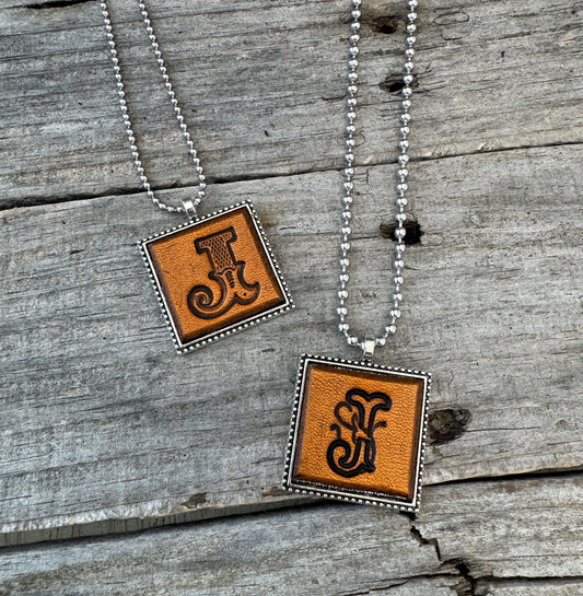 Two handmade Letter J leather initial necklaces showing Western Block and Floral Script lettering styles on warm brown leather pendants.