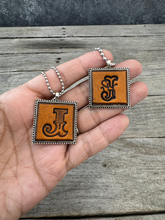 Western Block and Floral Script Letter J leather initial necklaces held in hand showing the size of the square silver-tone pendants.