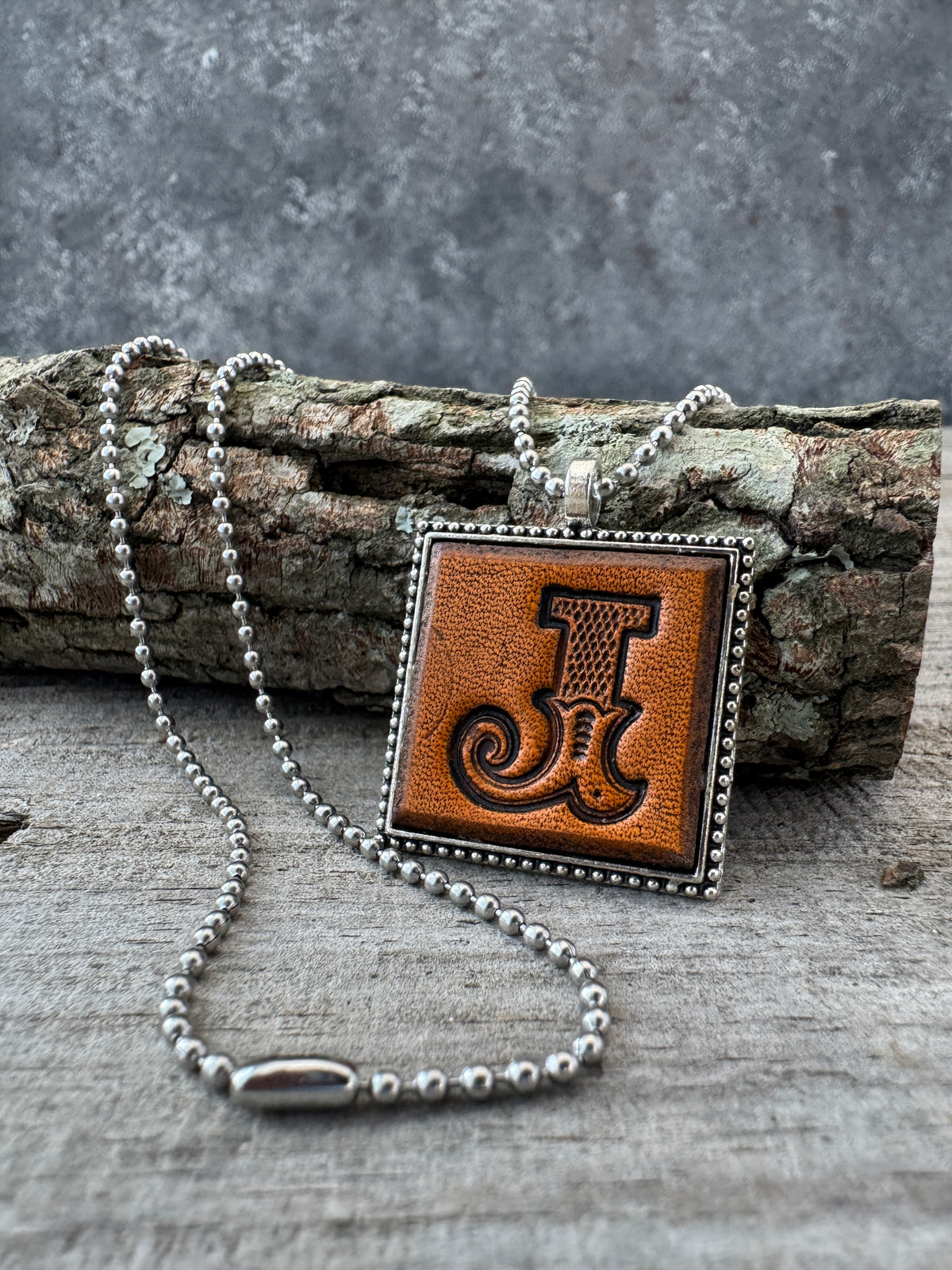 Hand-stamped Western Block Letter J leather necklace with silver-tone ball chain displayed against rustic tree bark and weathered wood.