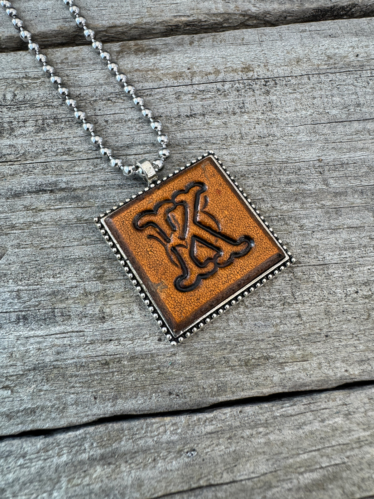 Handcrafted Letter K leather initial necklace featuring decorative Floral Script lettering stamped into warm brown leather with a silver-tone square pendant frame.
