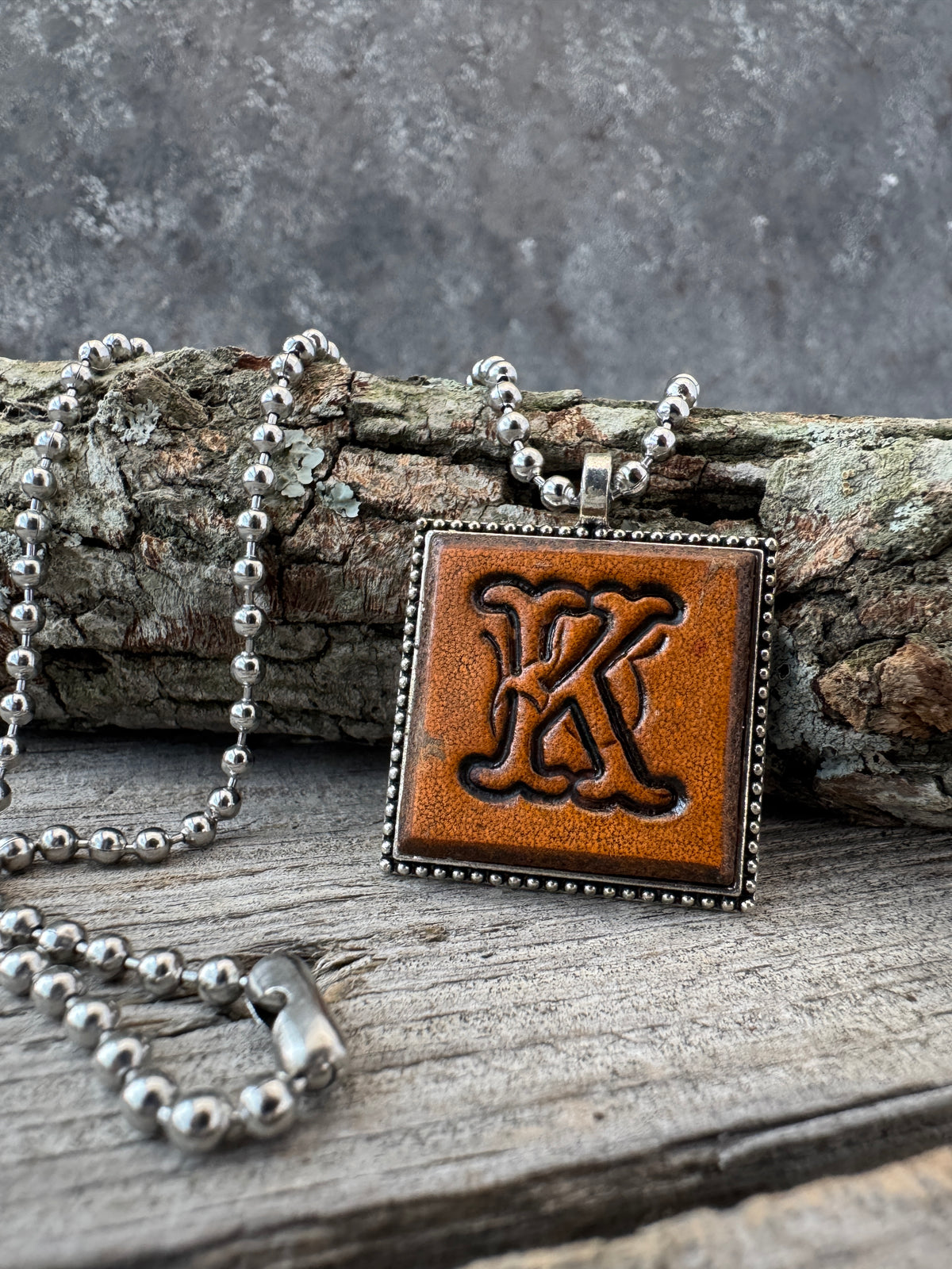 Floral Script Letter K leather pendant necklace with silver-tone ball chain displayed against natural tree bark and weathered wood.