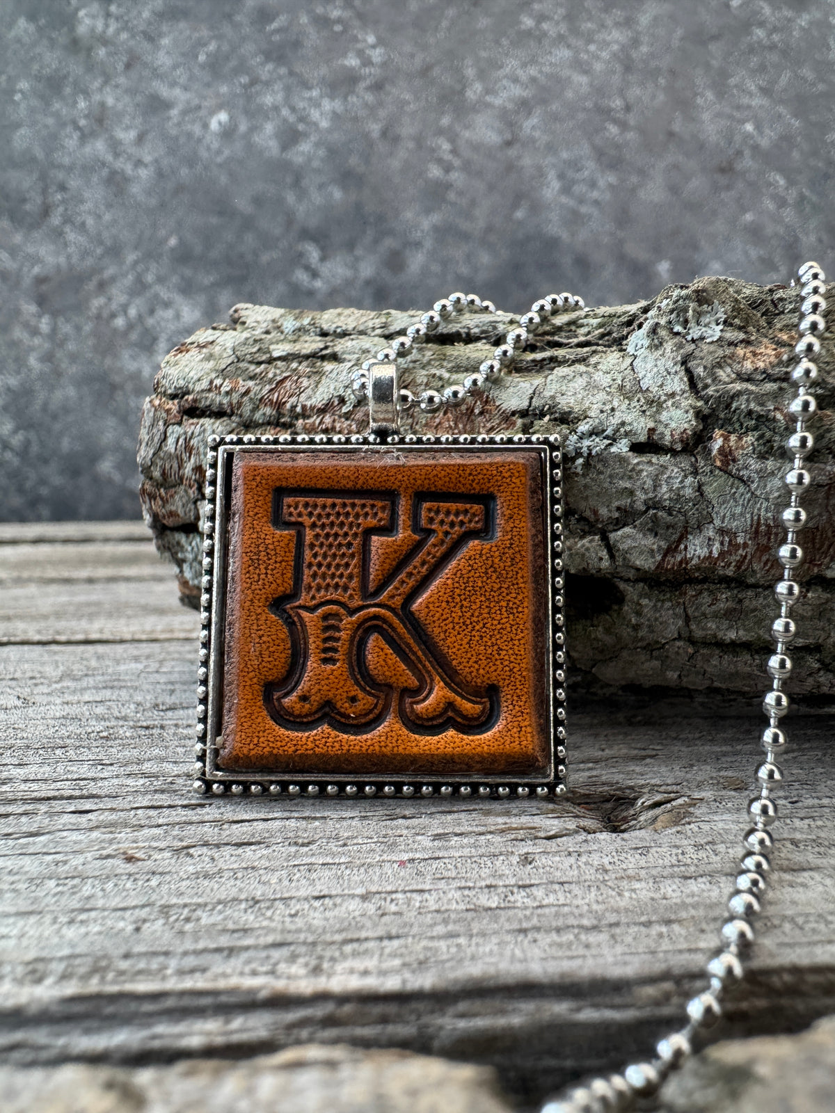 Hand-stamped Western Block Letter K leather necklace with silver-tone ball chain displayed against rustic tree bark and weathered wood.