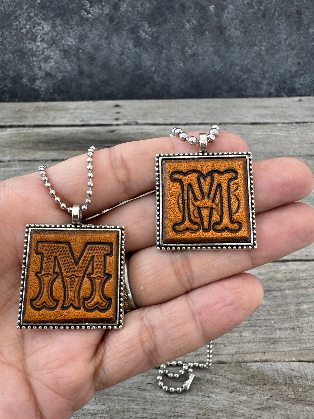 Western Block and Floral Script Letter M leather necklaces held in hand for size comparison