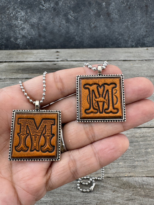 Western Block and Floral Script Letter M leather necklaces held in hand for size comparison