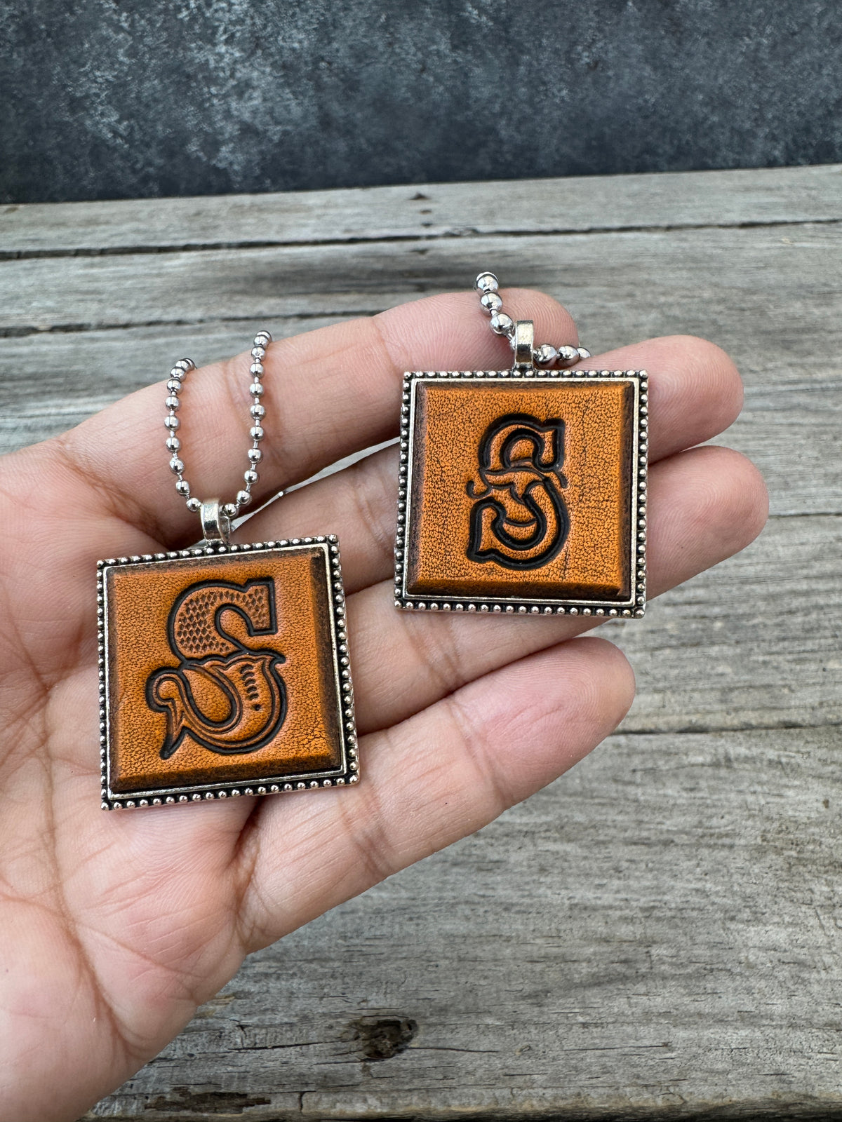 Western Block and Floral Script Letter S leather necklaces held in hand for size comparison