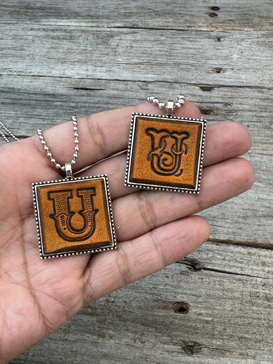 Western Block and Floral Script Letter U leather necklaces held in hand for size comparison
