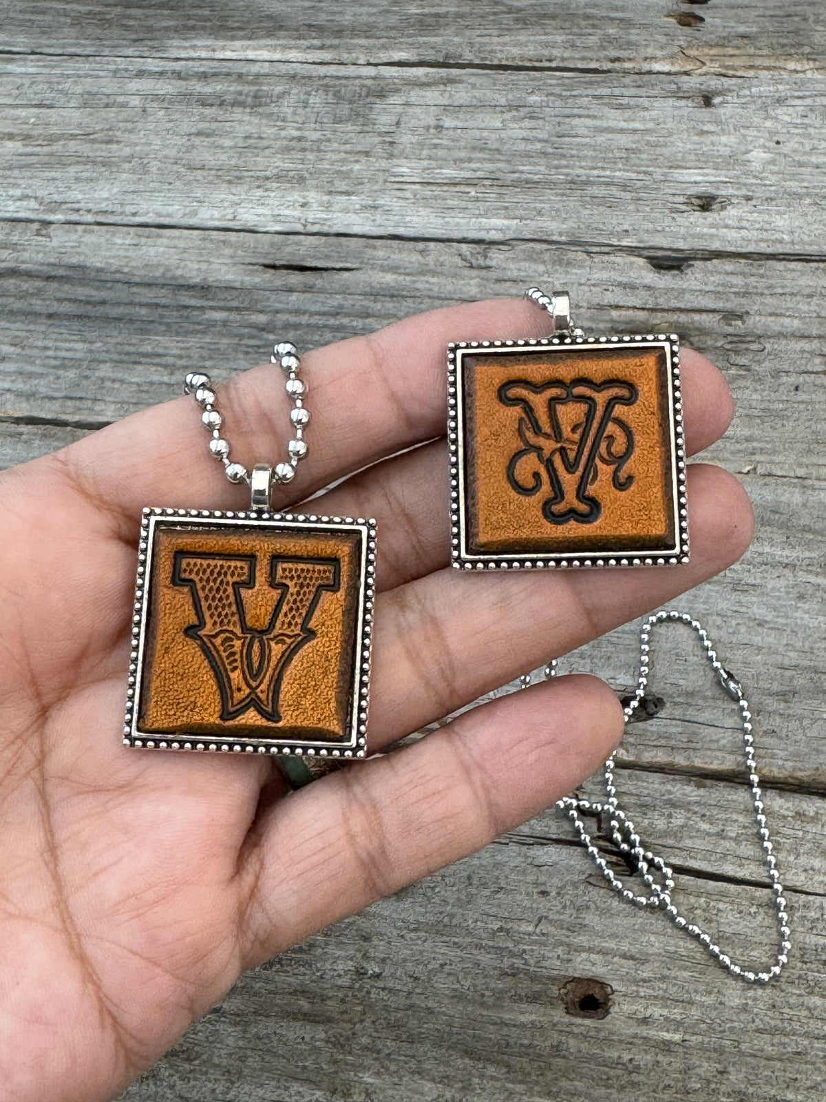 Western Block and Floral Script Letter V leather necklaces held in hand for size comparison