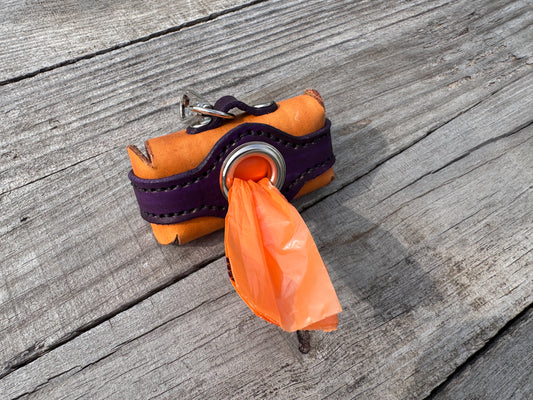 An orange leather dog poop bag holder with a purple strap and metal ring, holding an orange plastic bag, placed on a weathered gray wooden surface.