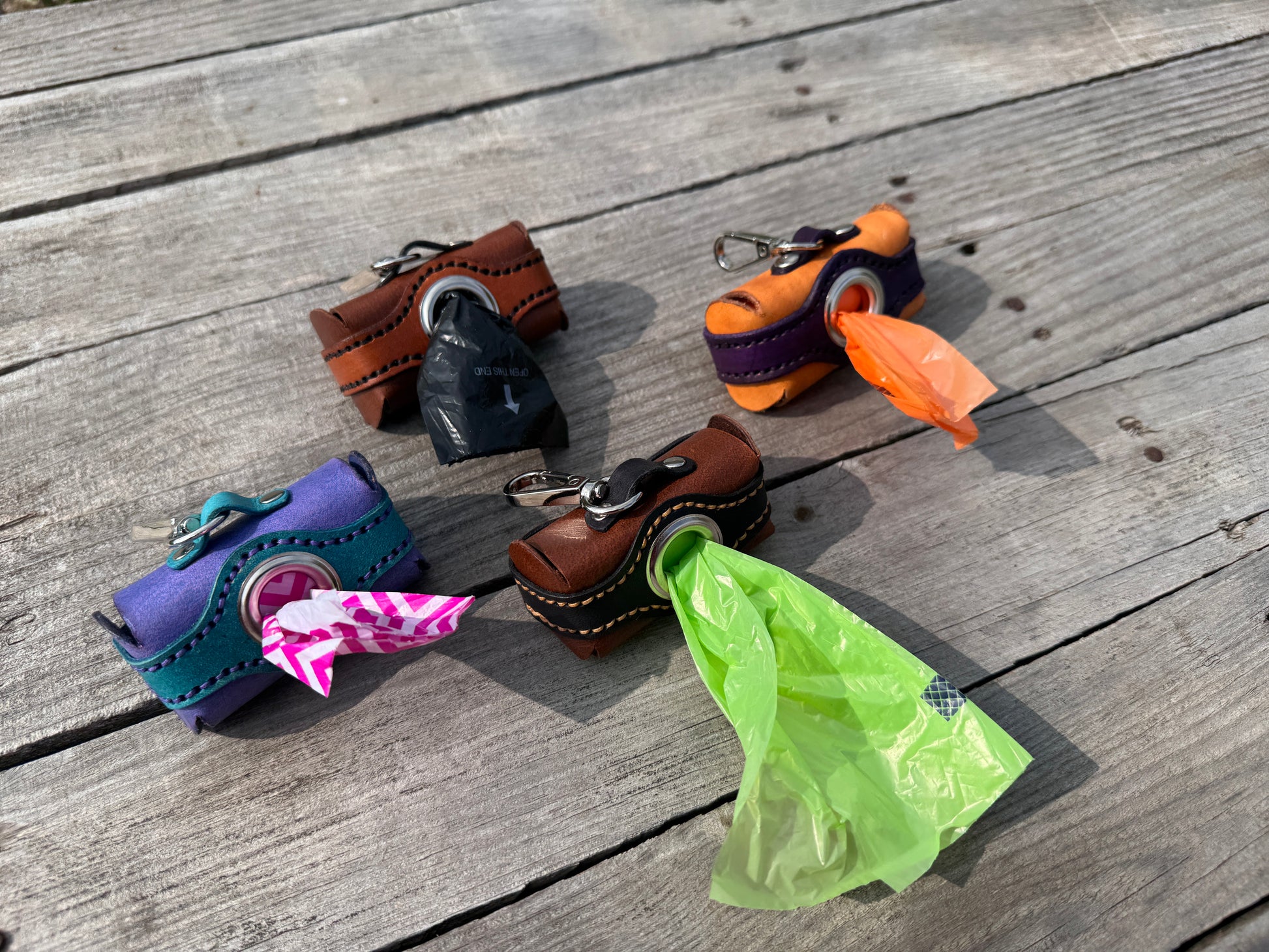 Four handmade leather dog poop bag holders, each with a different color combination and a plastic bag, arranged on a weathered wooden deck.
