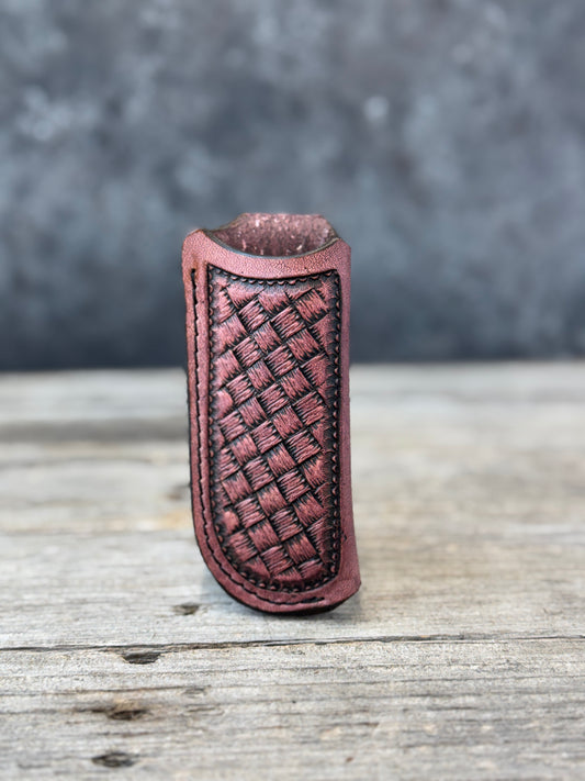 A reddish-brown custom stamped leather knife sheath with a woven basketweave pattern on the front, standing upright on a rustic wooden surface with a blurred gray background.