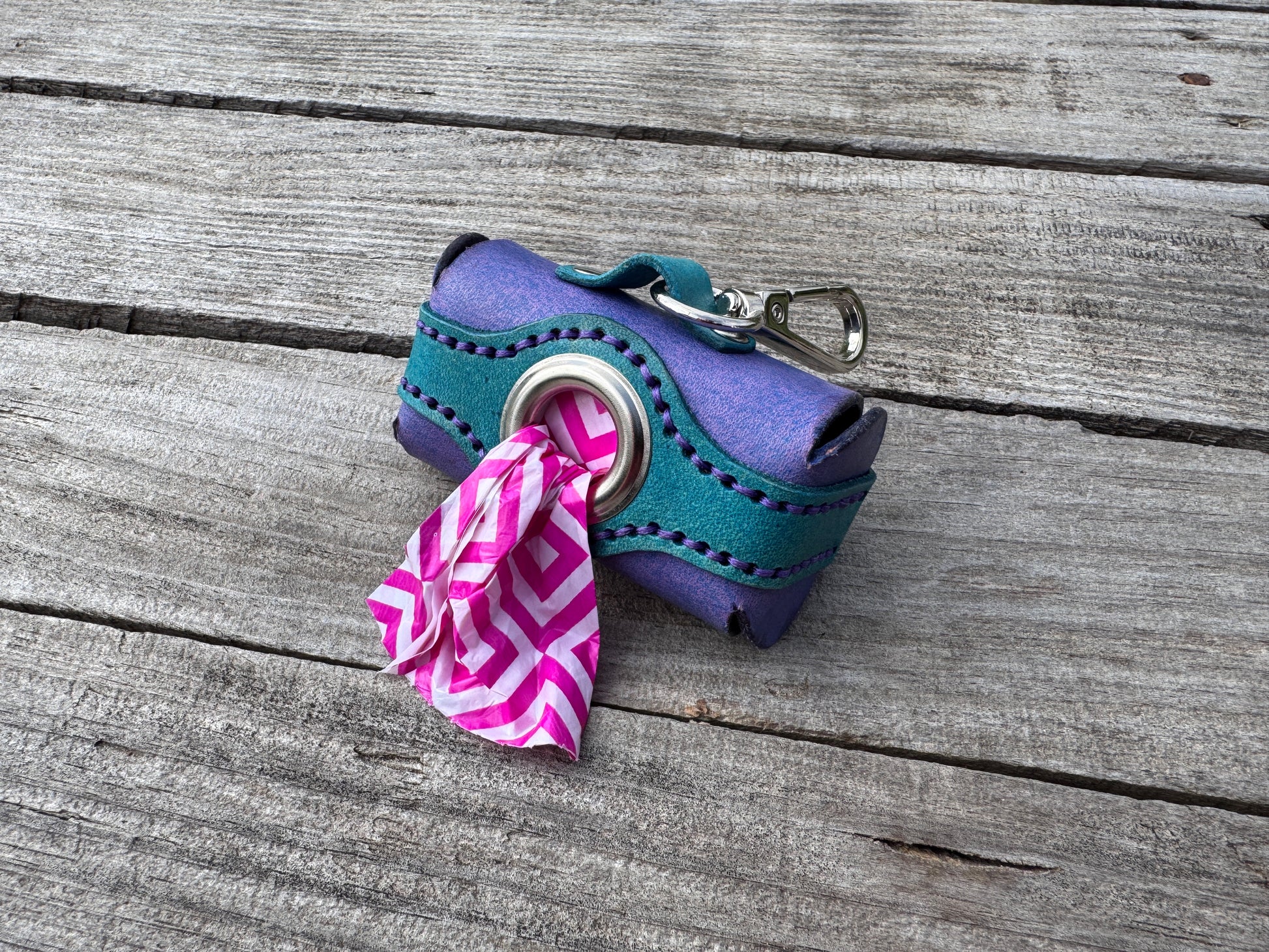 A small purple and teal dog leash accessory with a metal clip and a pink-and-white patterned poop bag, photographed on weathered gray wooden planks.