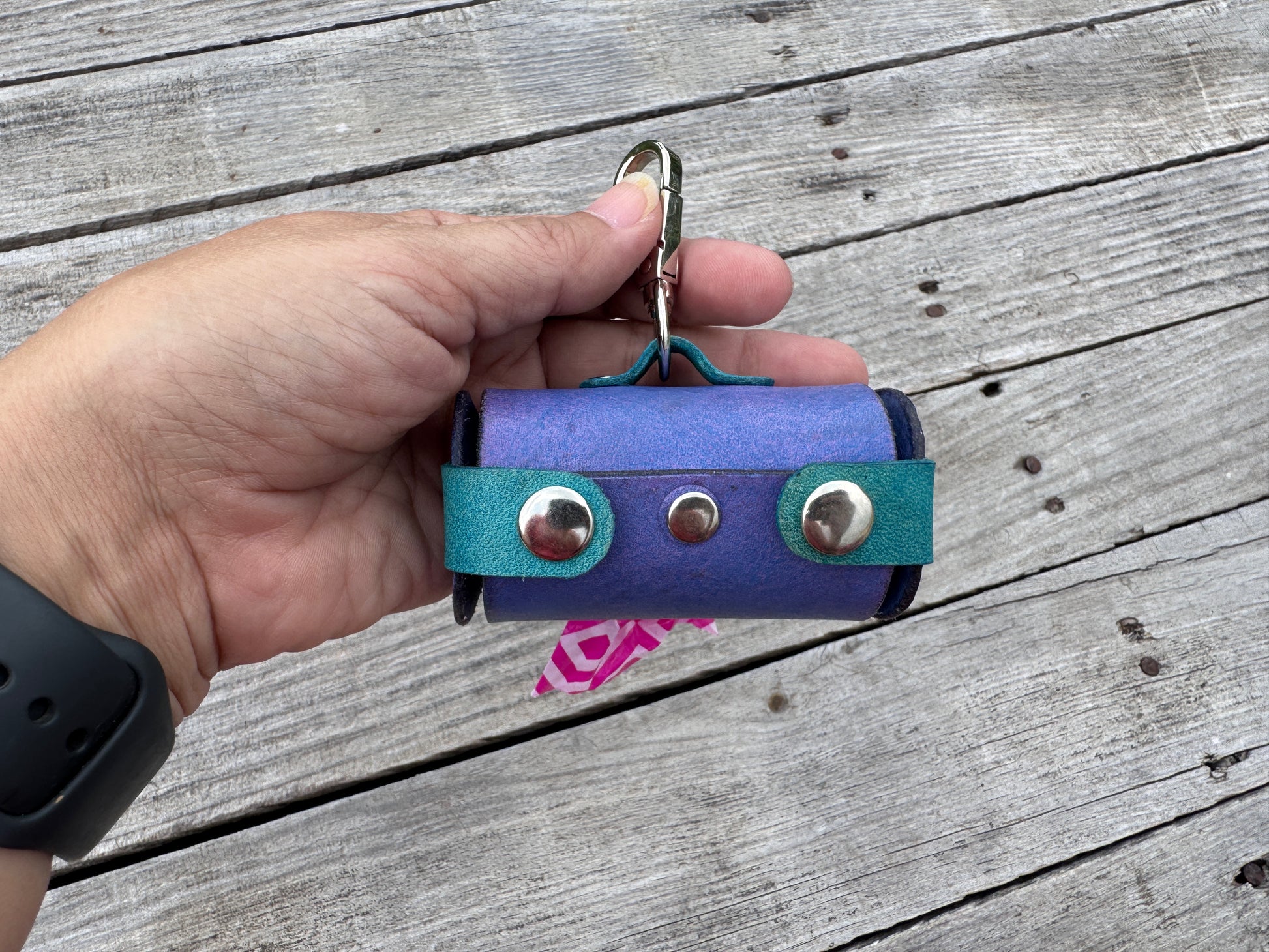 A hand holding a leather poop bag holder that is purple item with teal straps and silver-tone snaps, photographed outdoors against a weathered gray wooden plank surface.