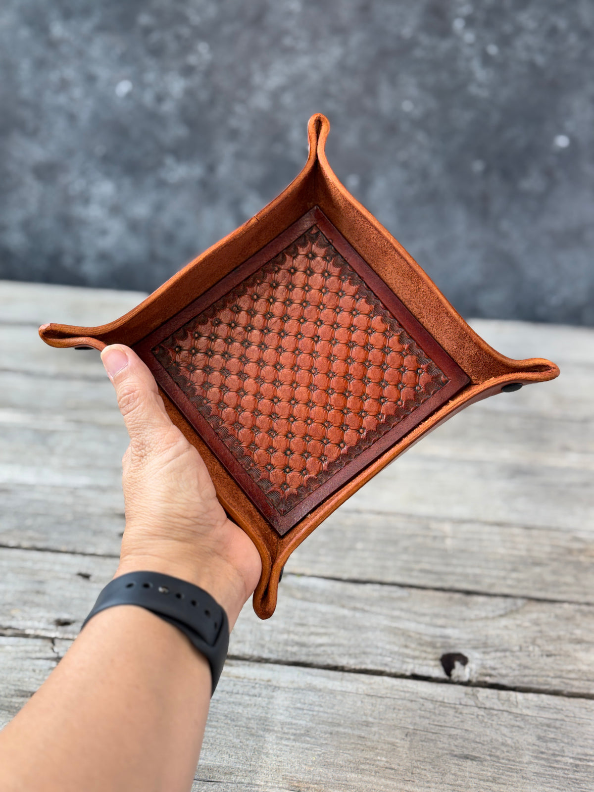Hand holding a woven brown leather valet tray on a wooden surface with a gray background