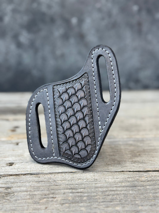 A steel grey stamped leather knife sheath with a textured, dragon scale-patterned center panel and grey contrast stitching, resting on a rustic wooden surface with a blurred gray background.