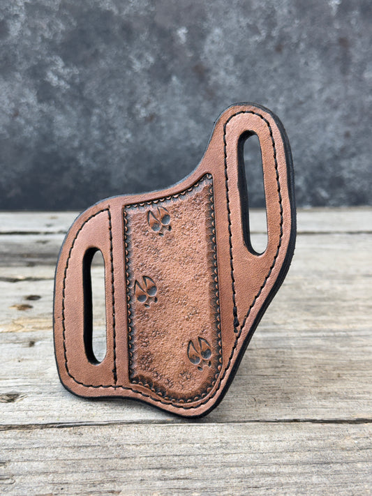 A custom leather knife sheath with cutout belt slots and a stamped deer track detail, photographed on a split gray and weathered wood background
