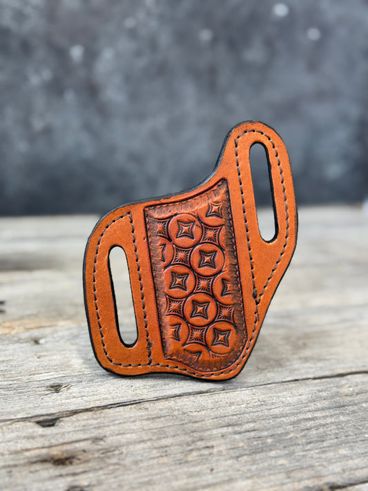 A custom leather knife sheath with a stamped geometric pattern, resting on a rustic wooden surface with a blurred gray background